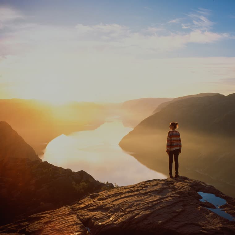 Kvinna står på ett högt berg och tittar ut över en dalgång i skymningsljus