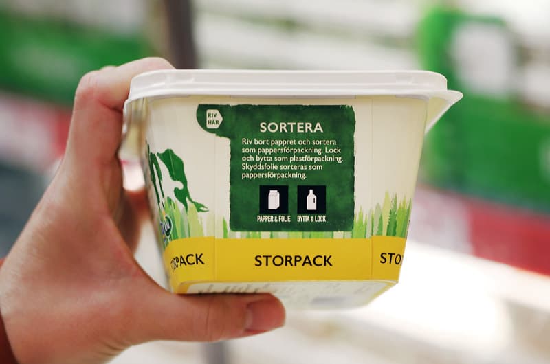 En hand som håller upp ett smörpaket med märkning som visar att förpackningen ska källsorteras som papper och plast.