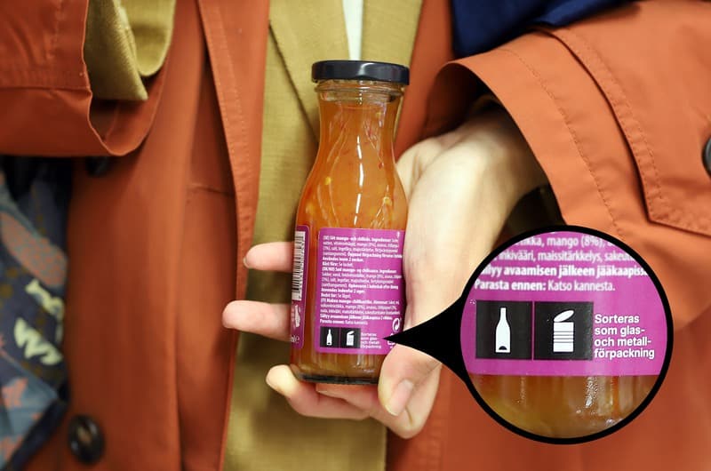 En hand som håller en flaska med mango- och chilisås med märkning som visar att förpackningen ska källsorteras som glas och metall.