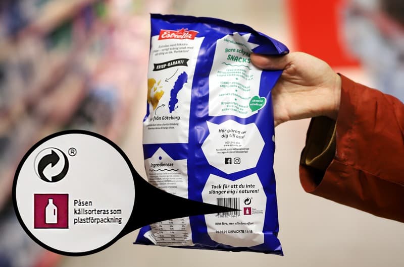 En hand som håller upp en chipspåse med märkning som visar att förpackningen ska källsorteras som plast.