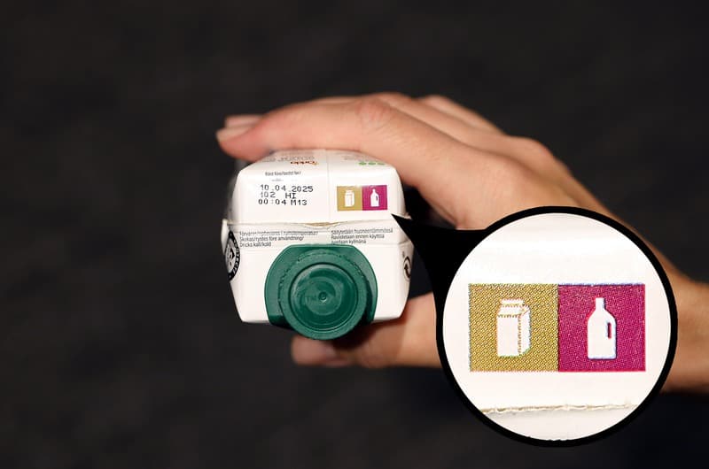 En hand som håller upp en dryckeskartong med märkning som visar att förpackningen ska källsorteras som papper och plast.