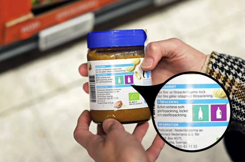 En hand som håller en jordnötssmörsburk med märkning som visar att förpackningen ska källsorteras som glas och plast.