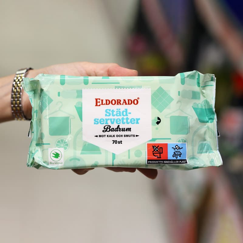 En hand som håller en förpackning med städservetter med märkning som visar att produkten innehåller plast.