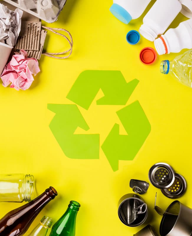 Förpackningar av olika materialslag mot en gul bakgrund. En grön återvinningssymbol ligger i mitten av bilden.